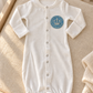 White Brit Milah baby gown with blue crown patch styled on ivory nursery linen