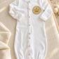 White Brit Milah crown patch baby gown styled on an ivory nursery linen surface
